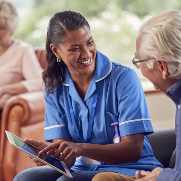Senior Couple At Home With Man Talking To Female Nurse Or Care Worker Using Digital Tablet