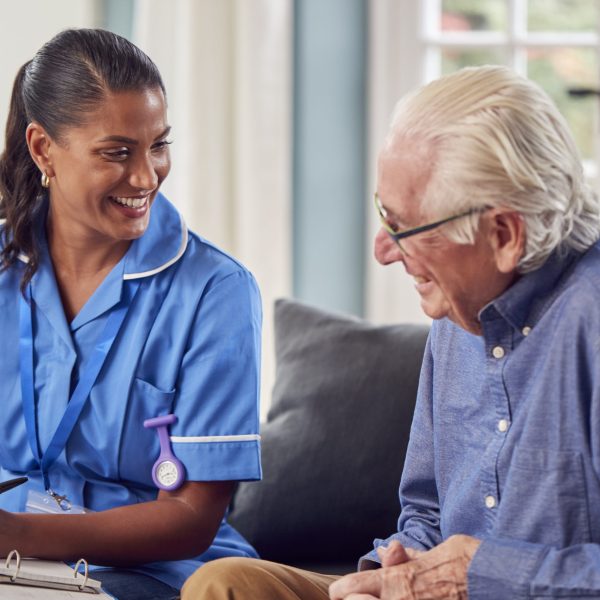 Senior Man At Home Talking To Female Nurse Or Care Worker In Uniform Making Notes In Folder