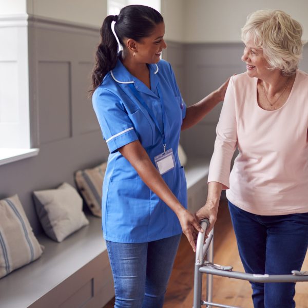 Senior Woman At Home Using Walking Frame Being Helped By Female Care Worker In Uniform
